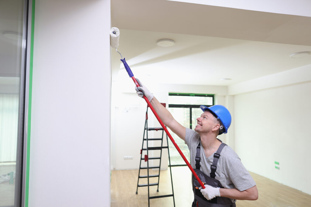 Portrait of smiling decorator in helmet painting wall with roller brush. Painting, renovation and Regular interior painting for commercial spaces