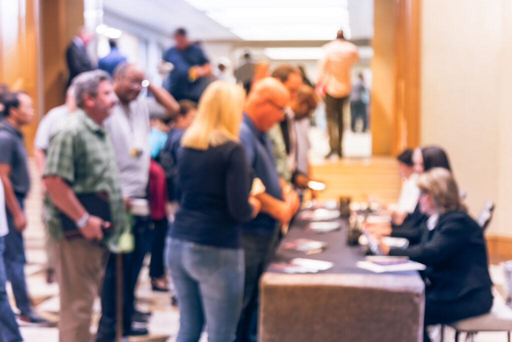 Blurred side view diverse participants at registration check-in workshop table. Multiethnic people check-in at hotel lobby near staircase and received instruction, event package from support staff